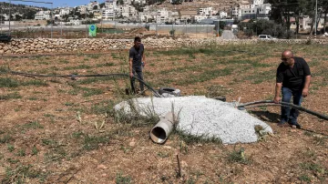 L'image montre deux hommes travaillant sur un terrain en plein air, probablement dans une zone rurale. Ils manipulent des tuyaux en plastique et se trouvent à proximité d'un tas de ciment ou de béton, ainsi qu'un petit tuyau qui sort du sol. À l'arrière-plan, on peut apercevoir des bâtiments et des collines. La végétation est clairsemée, et le sol semble fraîchement labouré ou non entretenu.