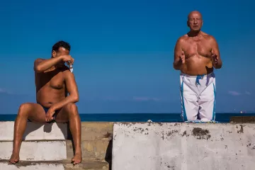 L'image montre deux hommes sur une structure en béton au bord de l'eau. L'un des hommes est assis, torse nu, avec les bras croisés sur un genou, semblant protéger ses yeux du soleil. L'autre homme, debout à côté de lui, est également torse nu et semble parler ou gesticuler, comme s'il était en pleine conversation. En arrière-plan, on peut voir un ciel bleu clair et une étendue de mer, créant une atmosphère estivale et décontractée.