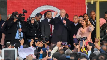 L'image montre un groupe de personnes rassemblées devant une scène où un homme, probablement un leader politique, prend la parole. Il est entouré de plusieurs personnes, dont des membres de la sécurité et des journalistes avec des caméras. La foule est très engagée, beaucoup de gens tiennent des téléphones pour filmer ou prendre des photos. L'arrière-plan présente des affiches ou des bannières, probablement en rapport avec un événement important. L'ambiance semble dynamique et pleine d'énergie.