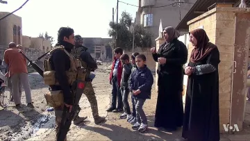تظهر في الصورة مجموعة من الأشخاص في شارع يبدو أنه في منطقة تتعرض للتوتر أو النزاع. في الجهة اليسرى، يوجد جندي مسلح يرتدي زيًا عسكريًا، ويتحدث مع مجموعة من النساء والأطفال. النساء يرتدين ملابس تقليدية، بينما يبدو الأطفال مهتمين بالمحادثة. خلفهم، يمكن رؤية مباني متضررة وأجواء تعكس حالة المنطقة.