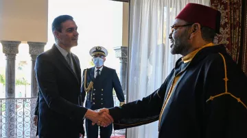 The image depicts a meeting between two men. One man, dressed in a suit, is shaking hands with the other man, who is wearing traditional attire, including a red fez. In the background, a military figure stands in uniform, observing the handshake. The setting appears to be an elegantly decorated room with large windows and elaborate architectural details.