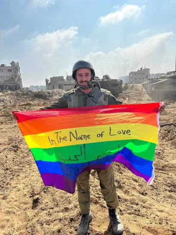 Un soldat souriant tient un drapeau arc-en-ciel sur un terrain dévasté.