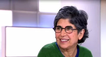 L'immagine mostra una donna con capelli grigi e ondulati, indossando occhiali e un maglione verde. Sembra sorridere e avere un'espressione accogliente. Sullo sfondo ci sono sfumature chiare che danno un'atmosfera luminosa e positiva.