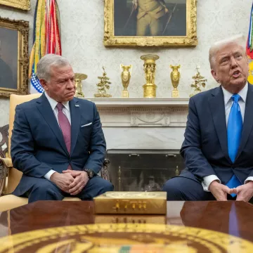 L'image montre deux hommes assis dans une pièce élégante, probablement dans un bureau officiel. L'un d'eux, portant un costume sombre, regarde avec une expression sérieuse, tandis que l'autre, en costume clair avec une cravate bleue, parle. Des drapeaux et des tableaux anciens décorent la pièce. Une table en bois est visible devant eux.