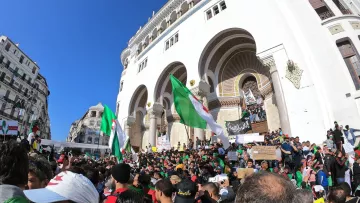 تظهر الصورة تجمعًا حاشدًا لمتظاهرين في مكان مفتوح، يحتشدون أمام مبنى تاريخي. يرفع المتظاهرون أعلام الجزائر بينما يحمل البعض لافتات تحمل شعارات. يتواجد عدد كبير من الأشخاص في الصورة، مما يبرز حماسهم ومشاركتهم الفاعلة. السماء زرقاء وظهور شعاع الشمس، مما يعكس أجواء نابضة بالحياة.