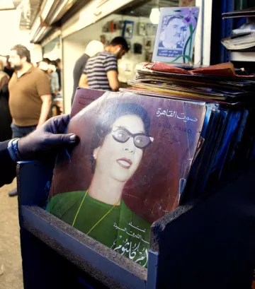 L'image montre une personne tenant un disque vinyle dans un marché. Le disque est de couleur foncée, avec une illustration d'une femme portant des lunettes et des vêtements vintage. En arrière-plan, on aperçoit d'autres personnes dans un environnement animé, probablement un marché ou une braderie, avec divers objets et éléments visibles autour. L'ambiance semble nostalgique, soulignant l'importance de la musique et de la culture visuelle.