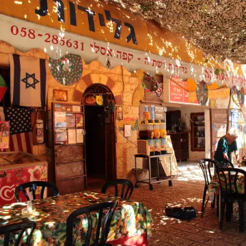 L'image montre un café ou une taverne en plein air, avec des tables et des chaises noires disposées sur un sol en pierre. Le lieu semble très accueillant, avec des nappes colorées sur les tables. Au-dessus, un toit en toile de camouflage laisse passer un peu de lumière. On peut voir des drapeaux accrochés aux murs, probablement des symboles de différentes nationalités. Des personnes assises discutent tranquillement dans cet environnement convivial. L'architecture est typique des bâtiments anciens, ajoutant une touche authentique à l'ambiance.