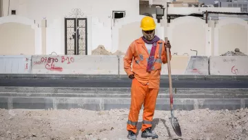 Nell'immagine si vede un lavoratore in un cantiere edile. Indossa una tuta arancione e un casco giallo per la sicurezza. Ha in mano una pala e sembra essere in pausa, osservando il terreno di fronte a lui. Sullo sfondo si possono notare alcune strutture e graffiti su un muro. L'ambiente appare polveroso e in fase di costruzione.