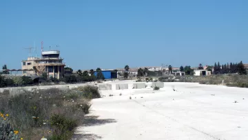 The image depicts an abandoned or derelict area, likely an old airstrip or airport. In the background, there is a control tower or building, which appears to be in disrepair. Surrounding the area are sparse patches of vegetation, including some wildflowers, and the ground is mostly composed of white gravel or concrete. The sky is clear and blue, indicating a bright, sunny day, and the overall scene conveys an atmosphere of neglect and desolation.