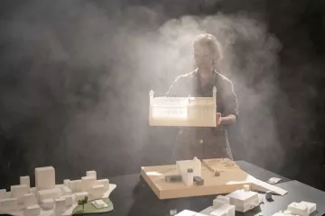 L'image montre une personne tenant une maquette d'un bâtiment, dans un environnement brumeux. Le fond est sombre, ce qui met en valeur la maquette et la silhouette de la personne. Sur la table, il y a d'autres maquettes et éléments architecturaux, suggérant une scène de travail ou de présentation liée à l'architecture. La lumière semble filtrer à travers la brume, créant une ambiance mystérieuse et artistique.