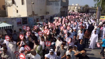 الصورة تظهر مجموعة كبيرة من الناس في مظاهرة أو احتجاج. المشاركون يحملون لافتات كبيرة عليها إشارات تعبر عن ممنوع أو خط أحمر. يرتدي الكثير منهم ملابس تقليدية، وتبدو الحشود منظمة. الخلفية تحتوي على مبانٍ وصور ملصقة على الجدران، مما يشير إلى أن الحدث يحدث في منطقة حضرية.