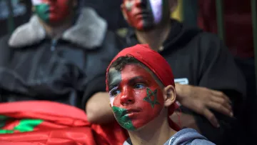 L'image montre un groupe de jeunes, dont un enfant au premier plan, qui ont le visage peint avec des motifs aux couleurs du drapeau marocain. L'enfant porte un bandana rouge et des dessins représentant le drapeau marocain et d'autres motifs sur son visage. En arrière-plan, deux autres personnes affichent également des visages peints, créant une atmosphère festive et engagée. L'ambiance semble liée à un événement de soutien ou à une célébration.