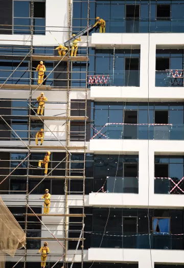 L'image montre un bâtiment en construction. On peut voir des échafaudages le long de la façade, sur lesquels plusieurs ouvriers vêtus de combinaisons jaunes et de casques de sécurité sont en train de travailler. Certains utilisent des outils pour peindre ou poser des éléments sur les fenêtres, tandis que d'autres montent et descendent sur les échafaudages. Le bâtiment a des fenêtres en verre, et des bandes de sécurité sont visibles autour de certaines zones. L'ensemble de la scène reflète un environnement de travail actif et organisé.