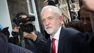 L'image montre un homme avec des cheveux gris et une barbe, vêtu d'un costume sombre et d'une cravate rouge. Il semble se déplacer dans un environnement urbain, entouré de journalistes et de caméras qui l'interrogent ou le photographient. L'atmosphère est animée et il est probable qu'il soit au centre de l'attention médiatique. Le cadre présente des bâtiments en briques, suggérant une ambiance citadine.