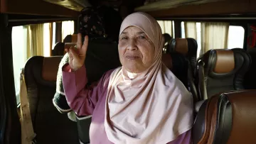 L'image présente une femme âgée à l'intérieur d'un bus. Elle porte un hijab de couleur claire et un vêtement de style traditionnel. Elle fait un signe de paix avec ses doigts tout en souriant. En arrière-plan, on peut apercevoir d'autres passagers et l'intérieur du bus avec des sièges confortables. L'atmosphère semble chaleureuse et amicale.