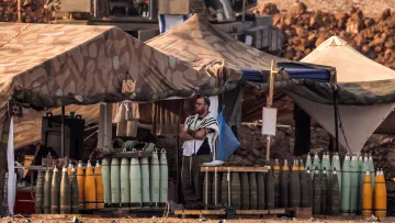 The image depicts a military encampment with tents set up in a rugged landscape. In the foreground, a soldier wearing a tallit (a traditional Jewish prayer shawl) stands near stacks of artillery shells arranged on a wooden platform. The tents are camouflaged, blending into the environment, and various military equipment is visible in the background. The setting conveys a sense of military readiness and activity.