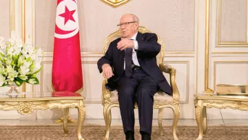 L'image montre un homme assis sur une chaise en or dans un environnement intérieur élégant. Il porte un costume sombre et des lunettes. À sa gauche, il y a un vase avec des fleurs blanches. Derrière lui, on aperçoit le drapeau tunisien. L'homme semble être en train de parler ou de gesticuler, indiquant une conversation ou une discussion en cours. Les murs et le décor environnant évoquent une atmosphère formelle et institutionnelle.