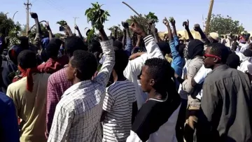 L'image montre un grand groupe de personnes rassemblées à l'extérieur, levez les bras en signe de protestation ou de soutien, tenant probablement des branches ou des plantes. Le paysage semble être rural, avec quelques bâtiments en arrière-plan et un ciel dégagé. L'ambiance semble dynamique et énergique, reflétant une mobilisation collective.