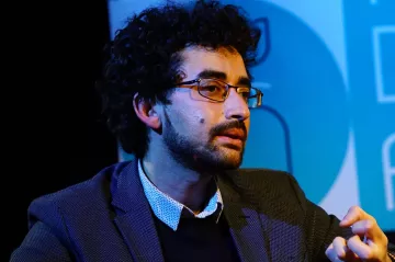 Homme avec des lunettes, cheveux bouclés, en discussion, vêtu d'une veste.