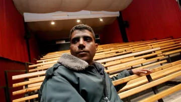 L'image montre un homme assis sur un banc dans un auditorium vide. L'homme porte une veste avec un col en fourrure et semble pensif, regardant vers l'avant. L'arrière-plan est composé de rangées de sièges en bois et de rideaux rouges, créant une atmosphère de théâtre ou de cinéma. La scène est bien éclairée, mettant en valeur l'homme et l'environnement autour de lui.