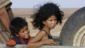 L'image montre deux enfants, un garçon et une fille, sur un véhicule. Le garçon, souriant, a les cheveux noirs et bouclés, et il semble joyeux. La fille, dont les cheveux sont également très volumineux, a une expression sérieuse ou pensive. Ils sont entourés d'un paysage désertique aux nuances de jaune et de beige. L'arrière-plan suggère un environnement rustique et chaud.