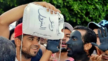 L'image montre un groupe de personnes en train de participer à une manifestation ou un rassemblement. Au premier plan, deux hommes attirent l'attention. L'un d'eux porte un récipient en plastique sur la tête, tandis que l'autre a le visage peint en noir. Ils semblent discuter ou se divertir, entourés de plusieurs autres personnes qui participent à l'événement. L'ambiance paraît animée et engagée, mettant en avant des expressions de solidarité ou de protestation.