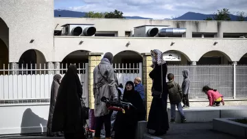 The image depicts a scene outside a building with several people present. Some individuals are wearing traditional clothing, including hijabs, while others are dressed in more casual attire. In the background, there are air conditioning units on the building, and a few trees and mountains can be seen in the distance. The setting appears to be a community space or public area, with a mix of adults and children interacting with one another. The atmosphere seems calm and social.