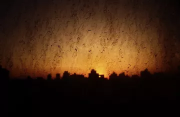 L'image montre un paysage urbain au crépuscule, avec des silhouettes de bâtiments se découpant contre un ciel orangé. La texture de la vitre est visible, ajoutant une dimension bruitée et floue à la scène. Les reflets et les imperfections de la vitre créent une atmosphère légèrement mystérieuse, tandis que la lumière du soleil couchant rappelle la fin de la journée.