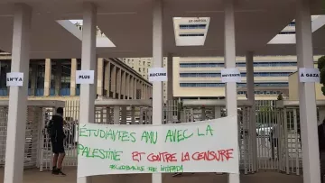 L'image montre l'entrée d'Aix Marseille Université, avec des panneaux affichant des messages. Au premier plan, il y a une grande bannière sur laquelle est écrit en rouge et en vert : "Étudiantes AMU avec la Palestine et contre la censure". L'atmosphère semble refléter un soutien à la cause palestinienne et une dénonciation de la censure. Quelques personnes sont visibles dans la scène, et le cadre est universitaire.