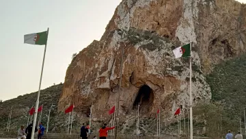 L'image montre une imposante falaise avec une grande ouverture, probablement une grotte, qui se trouve à sa base. Des drapeaux algériens flottent autour, ajoutant une touche de couleur au paysage. Au premier plan, on peut voir des personnes marchant près des drapeaux. Le ciel est teinté de lumière douce, suggérant peut-être un lever ou un coucher de soleil. L'ensemble évoque un lieu naturel remarquable avec une signification culturelle ou historique.