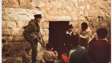 L'image montre un groupe de personnes près d'une porte en pierre. À gauche, un soldat armé en uniforme vert se tient debout, gardant l'entrée. Deux hommes adultes discutent, tandis qu'un enfant et un autre homme semblent observer la scène. L'environnement est marqué par des murs de pierre brute, suggérant un endroit ancien ou historique. L'atmosphère paraît chargée, et les expressions des personnes indiquent une interaction sérieuse.