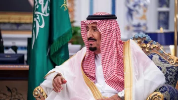 The image shows a man seated in a lavish decor room, wearing traditional attire that includes a white thobe and a red and white checked keffiyeh held in place by an agal. He has a thoughtful expression and is gesturing with his hand. Behind him, there is a green flag, likely representing Saudi Arabia, and opulent furnishings in the background.