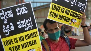 The image depicts a person holding two large signs that read "LOVE IS NO CRIME! STOP HATRED!" in both English and Kannada, a language spoken in India. The individual is wearing a face mask and appears to be participating in a demonstration or rally, advocating for love and against hate. The atmosphere suggests a sense of activism and a call for social change, with a crowd visible in the background. The signs are bright and eye-catching, emphasizing the message strongly.