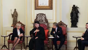 L'image montre une scène d'une réunion ou d'un entretien formel. Trois personnes sont assises dans des fauteuils ornés, au milieu d'une pièce décorée de manière élégante. L'une des personnes, portant une tenue ecclésiastique, semble être en train de parler, tandis que les autres l'écoutent attentivement. Les murs sont ornés de statues et d'œuvres d'art, ce qui suggère un environnement culturel et riche en histoire.