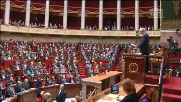 L'image montre une scène à l'Assemblée nationale, un lieu de débat politique en France. On y voit un grand amphithéâtre avec de nombreux sièges, occupés par des députés. Au premier plan, un homme se tient debout devant un pupitre, probablement en train de faire un discours ou de présenter une proposition. Les députés semblent attentifs et sont disposés en plusieurs rangées, tandis que certains sont assis à des tables sur le côté. L'architecture est typiquement institutionnelle, avec des éléments ornementaux et une atmosphère solennelle.