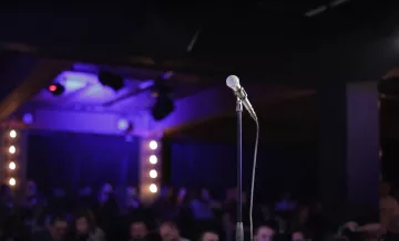 L'image montre un microphone sur un pied, au premier plan, sur une scène sombre. À l'arrière-plan, on aperçoit une audience floue et des lumières tamisées qui créent une ambiance de spectacle.