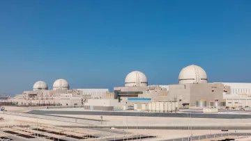 La imagen muestra una planta de energía nuclear con varias estructuras de reactor. Hay tres domos visibles en la parte superior de los reactores, que son características típicas de las centrales nucleares. La planta parece moderna y está rodeada de un entorno desértico, con un cielo claro y azul en el fondo. Hay edificios de soporte y áreas de estacionamiento en primer plano, lo que sugiere que es una instalación grande y compleja.