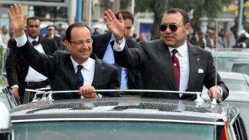 L'image montre deux hommes qui semblent être dans une voiture décapotable. L'un d'eux, à gauche, porte un costume sombre avec une cravate, tandis que l'autre à droite porte un costume avec des accessoires distinctifs. Ils saluent la foule avec la main, semblant sourire et être en bonne humeur. En arrière-plan, il y a des personnes et des voitures, probablement lors d'un événement officiel ou d'une visite. Le temps semble être pluvieux, avec des gouttes d'eau visibles sur la voiture.