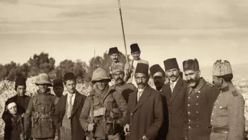 The image depicts a group of men dressed in historical military and civilian attire from an earlier era. In the foreground, a prominent figure holding a white flag stands among a diverse group of soldiers and civilians. The background features a rugged landscape, typical of a battlefield or a military campaign setting. The expressions and postures of the individuals suggest a moment of significance, possibly a truce or a ceremony. Overall, the image captures a poignant moment in history, highlighting the personalities and attire of the time.