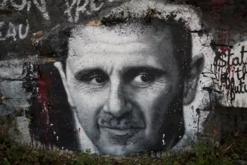 L'image montre un graffiti représentant un visage en noir et blanc sur un mur en pierre. Les traits du visage sont bien définis, avec un regard intense et expressif. Le fond est partiellement recouvert de peinture, et on peut voir des éclaboussures de couleur rouge, ajoutant une dimension dramatique à l'œuvre. Ce type d'art urbain est souvent utilisé pour faire passer des messages ou des commentaires sociaux.