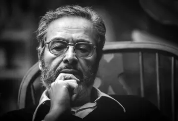 L'image montre un homme assis sur une chaise, arborant une barbe et des lunettes. Il semble profond dans ses pensées, avec une main sous le menton. L'éclairage donne un aspect sérieux à son expression, et le fond flou met en avant son visage. L'atmosphère est introspective et contemplative.