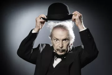 L'image montre un homme âgé portant un costume noir avec un nœud papillon. Il tient un chapeau melon avec une main, et son autre main semble légèrement levée. Ses cheveux sont ébouriffés, ce qui donne un aspect excentrique à son expression. L'arrière-plan est sobre, avec une lumière qui crée un effet dramatique, mettant en valeur son visage et son chapeau.