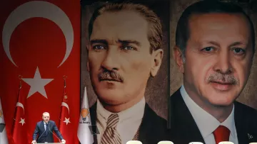 La imagen muestra a un orador frente a una gran bandera de Turquía, con retratos de dos figuras prominentes en la historia del país. A la izquierda, está el retrato de Mustafa Kemal Atatürk, el fundador de la República de Turquía, y a la derecha, el retrato del actual presidente, Recep Tayyip Erdoğan. El fondo es rojo, con símbolos nacionales y banderas que complementan el escenario, sugiriendo un evento político significativo.