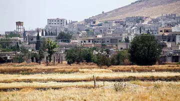 L'image montre un paysage rural avec des bâtiments en arrière-plan, probablement une petite ville ou un village. Au premier plan, il y a des champs avec de l'herbe sèche ou des cultures en attente de récolte. On peut voir une colline à l'arrière-plan, ainsi qu'un réservoir d'eau. La végétation est présente, avec quelques arbres éparpillés. L'atmosphère semble calme et ensoleillée, dégageant une ambiance de milieu rural.