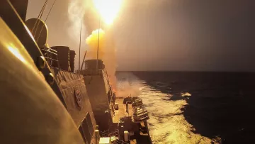 L'immagine mostra una scena marittima in cui un missile viene lanciato da una nave da guerra. Si vede il missile che viene sparato con un forte bagliore e un'esplosione di fumi dietro di esso. In primo piano si possono notare le strutture della nave e il mare in movimento, evidenziato da schiuma bianca. Il cielo appare velato e si intuisce un momento di grande intensità e potenza militare.