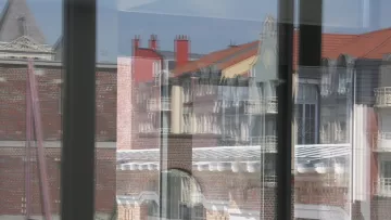 این تصویر نمایی از ساختمان‌ها را از طریق شیشه‌های یک ساختمان دیگر نشان می‌دهد. شیشه‌ها باعث ایجاد انعکاس‌هایی از بیرون می‌شوند که موجب می‌شود جزئیات معماری و رنگ‌های دیوارها و بام‌ها به خوبی نمایان شود. در پس‌زمینه، سقف‌های شیب‌دار و ساختمان‌های مختلف با طراحی‌های متفاوت دیده می‌شود، و نور آفتاب به تصویر جلوه‌ای جذاب می‌بخشد. فضای کلی تصویر حالت مدرنی دارد در حالی که عناصر تاریخی نیز در آن قابل مشاهده است.