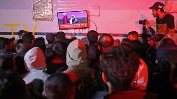 L'image montre une foule de personnes rassemblées, observant attentivement un écran au-dessus d'eux. L'écran affiche un discours ou une annonce, probablement en rapport avec des événements d'actualité. Certaines personnes dans la foule portent des capuches et des vêtements chauds, ce qui suggère qu'il fait frais. Un individu à droite prend des photos, ajoutant une dimension journalistique à la scène. L'éclairage est tamisé, créant une ambiance particulière.