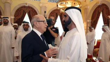 The image depicts two men engaging in conversation, likely in a formal setting. One man is dressed in a dark suit and glasses, while the other is in traditional Emirati attire, including a white kandura and headscarf. They appear to be having a cordial exchange, with the background showing a lavishly decorated room, including red curtains, ornate furniture, and a gathering of individuals in similar traditional attire. The atmosphere seems formal and respectful.