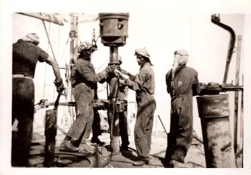 L'image représente un groupe de travailleurs en train de manipuler un équipement de forage, vraisemblablement sur un chantier pétrolier ou minier. Les ouvriers portent des vêtements de travail et des turbans, probablement pour se protéger de la poussière ou de la chaleur. Ils semblent concentrés sur leur tâche, travaillant ensemble sur une machine visible au centre de l'image. L'ambiance paraît industrialisée et intense, typique des travaux de forage au début du XXe siècle.