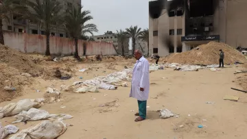 L'image montre un homme en blouse blanche se tenant debout sur un terrain désertique, entouré de sacs ressemblant à des corps. En arrière-plan, on peut voir des bâtiments endommagés et des morceaux de débris éparpillés sur le sol. La scène évoque un environnement de désastre, avec une atmosphère sombre et préoccupante, suggérant des événements tragiques. Des palmiers se dressent également sur le site, ajoutant un contraste avec la gravité de la situation.